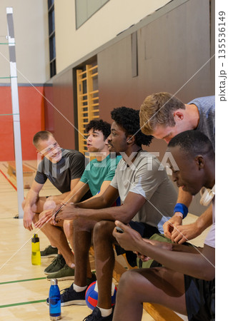 Diverse male volleyball team sitting, chatting in sports hall on bench with water bottles 135536149