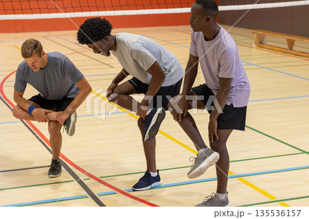 Diverse male teammates performing quadriceps stretches on gym volleyball court with net, bench 135536157