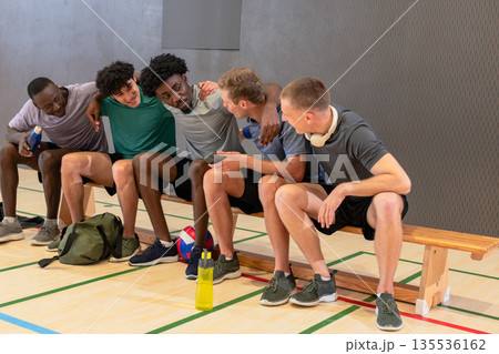 Diverse male partners sitting on bench at gym, chatting after volleyball game with water bottles Diverse male partners sitting on bench at gym, chatting after volleyball game with water bottles 135536162
