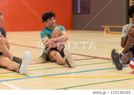 Diverse male teammates sitting on gym floor stretching legs by volleyball and bench 135536165