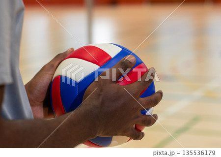 African american male athlete gripping red white, blue volleyball on gym court with net post African american male athlete gripping red white, blue volleyball on gym court with net post 135536179