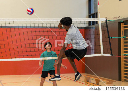 Diverse male friends rallying volleyball over net at school gym with bench, climbing bars Diverse male friends rallying volleyball over net at school gym with bench, climbing bars 135536188