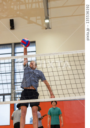 Diverse male teammates spiking volleyball at net in gym with red panels and overhead light Diverse male teammates spiking volleyball at net in gym with red panels and overhead light 135536192