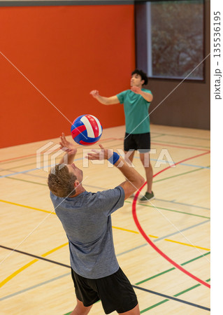 Adult male partners raising arms to set volleyball on gym court with painted lines windowed panels Adult male partners raising arms to set volleyball on gym court with painted lines windowed panels 135536195