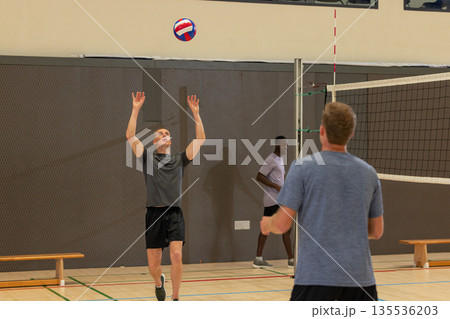 Diverse male friends raising hands, sending red white blue volleyball over net in gymnasium Diverse male friends raising hands, sending red white blue volleyball over net in gymnasium 135536203