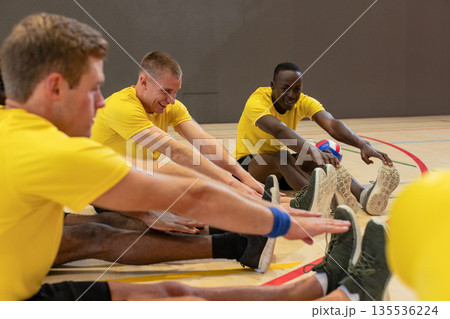 Male volleyball teammates in yellow shirts stretching legs on gym court holding volleyball 135536224