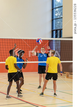 Diverse male volleyball athletes in yellow and blue shirts jumping at net chasing ball in gymnasium Diverse male volleyball athletes in yellow and blue shirts jumping at net chasing ball in gymnasium 135536228