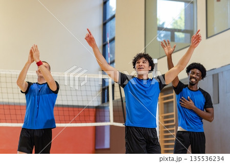 Diverse male volleyball athletes celebrating point in gym at volleyball net in blue black jerseys 135536234