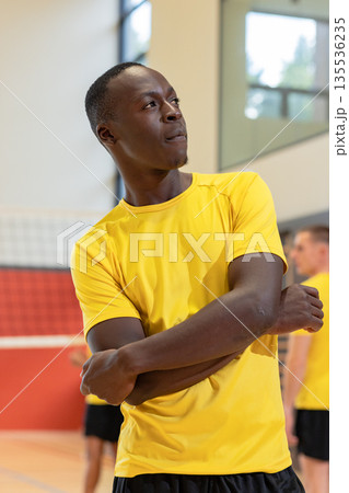 African american male player crossing arms by volleyball net in gym wearing yellow jersey shorts 135536235