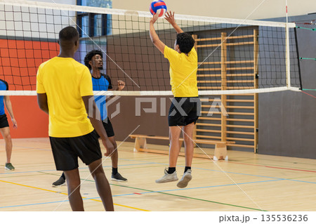 Diverse male volleyball players leaping and blocking on gym court with net, red-white-blue ball 135536236