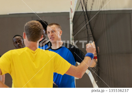 Diverse male volleyball athletes wearing wristband clasping hands through net at gym 135536237