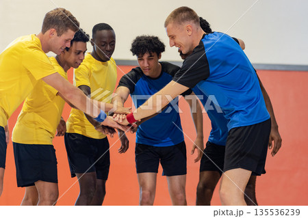 Diverse male athletes huddling placing hands at center in gymnasium in yellow and blue jerseys Diverse male athletes huddling placing hands at center in gymnasium in yellow and blue jerseys 135536239