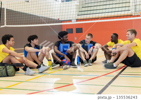 Diverse male partners sitting on gym court under net with volleyball, water bottles and sports bag 135536243
