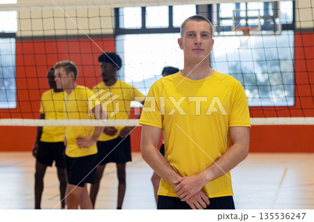 Diverse male partners in yellow jerseys and shorts standing on volleyball court in gym with net 135536247