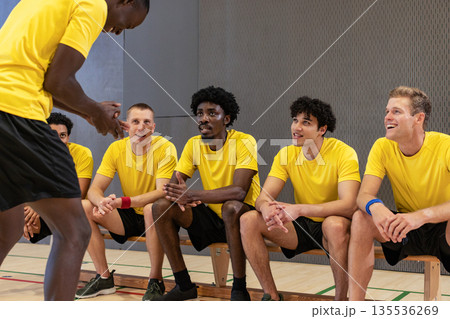Diverse male teammates talking around wooden bench in gymnasium with court lines, yellow jerseys Diverse male teammates talking around wooden bench in gymnasium with court lines, yellow jerseys 135536269