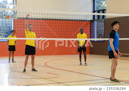 Diverse male volleyball athletes jumping and blocking on gym court with net, lines 135536284