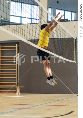 Male volleyball player jumping at net inside sports hall wearing yellow shirt, black shorts 135536285