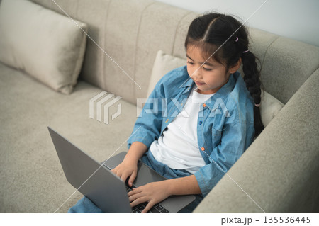 Young girl focused on laptop while sitting on a cozy couch with a calm atmosphere, engaging in online learning or creative activities at home Young girl focused on laptop while sitting on a cozy couch with a calm atmosphere, engaging in online learning or creative activities at home 135536445