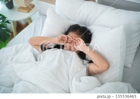 Young Girl Rubbing Eyes in Bed Surrounded by Cozy White Blankets and Pillows on a Calm Morning, Capturing Sleepy Moments and Household Life 135536465