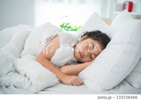 Young girl peacefully sleeping on a cozy bed surrounded by soft pillows in a bright and airy room, emphasizing calm and comfort in a serene setting. 135536466
