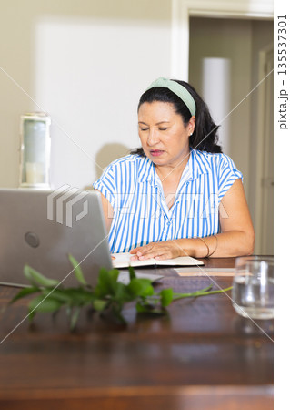Senior woman working on laptop at home, writing notes and concentrating 135537301