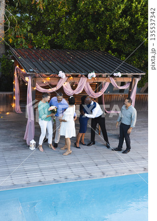 Exchanging hugs and flowers, diverse group celebrating by poolside under pergola Exchanging hugs and flowers, diverse group celebrating by poolside under pergola 135537342