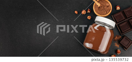 Chocolate spread in glass jar with cocoa powder and chocolate pieces on black background with copy space 135539732