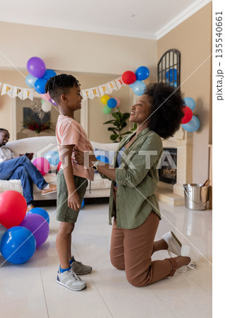 Mother kneeling, smiling at son in living room decorated with colorful balloons Mother kneeling, smiling at son in living room decorated with colorful balloons 135540661