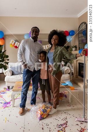 Family celebrating birthday at home, smiling with balloons and confetti on floor Family celebrating birthday at home, smiling with balloons and confetti on floor 135540715