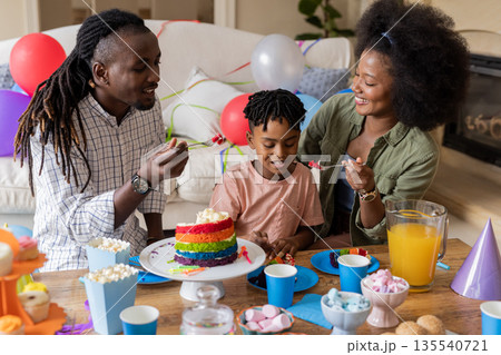 Celebrating birthday, parents and son enjoying rainbow cake and snacks at home 135540721