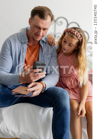 Father and daughter sitting on bed, looking at smartphone together, smiling 135540740