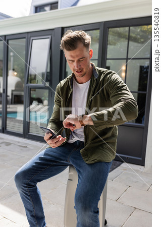 Man checking time on watch while holding smartphone outside modern home Man checking time on watch while holding smartphone outside modern home 135540819