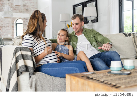 Family relaxing on sofa, young girl touching pregnant mother's belly, smiling warmly Family relaxing on sofa, young girl touching pregnant mother's belly, smiling warmly 135540895