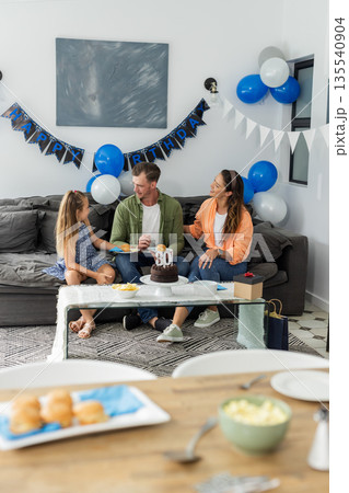 Celebrating birthday, family sitting on sofa with cake and decorations at home Celebrating birthday, family sitting on sofa with cake and decorations at home 135540904