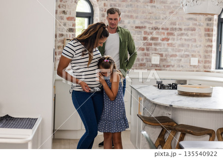 Expectant mother and daughter embracing in kitchen while father watches lovingly Expectant mother and daughter embracing in kitchen while father watches lovingly 135540922
