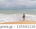 Young woman standing barefoot at shoreline facing calm ocean waves, wearing straw hat and backpack, travel and freedom concept with wide horizon  135541110