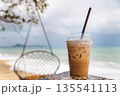 Iced coffee in plastic cup on beach table with ocean waves and hanging chair in background, tropical cafe mood, summer refreshment 135541113