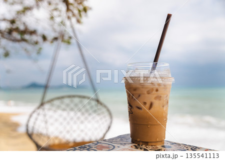 Iced coffee in plastic cup on beach table with ocean waves and hanging chair in background, tropical cafe mood, summer refreshment Iced coffee in plastic cup on beach table with ocean waves and hanging chair in background, tropical cafe mood, summer refreshment 135541113