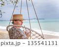 Middle aged woman relaxing in hanging chair on tropical beach, holding cold drink, ocean waves and cloudy sky in background, calm travel lifestyle  135541131