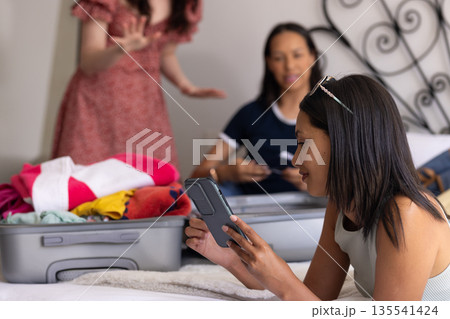 Packing suitcase on bed, young women with diverse backgrounds chat happily Packing suitcase on bed, young women with diverse backgrounds chat happily 135541424