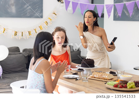 Celebrating birthday, three women gather around table with chocolate cake Celebrating birthday, three women gather around table with chocolate cake 135541533