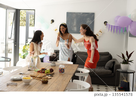 Laughing and celebrating, three young women gather around birthday cake at home Laughing and celebrating, three young women gather around birthday cake at home 135541578