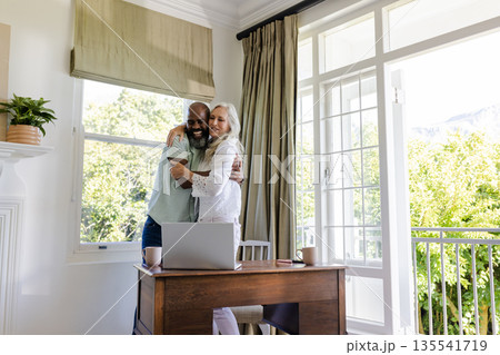 Couple embracing joyfully near desk with laptop, sunlight streaming through window 135541719