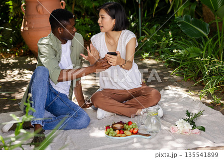 Sharing conversation and holding smartphone, couple having picnic outdoors together Sharing conversation and holding smartphone, couple having picnic outdoors together 135541899
