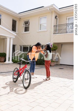 African American mother helping daughter with helmet near bicycle outside home 135541993