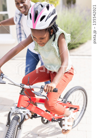 Father supporting African American child riding bicycle outdoors, both smiling 135542021