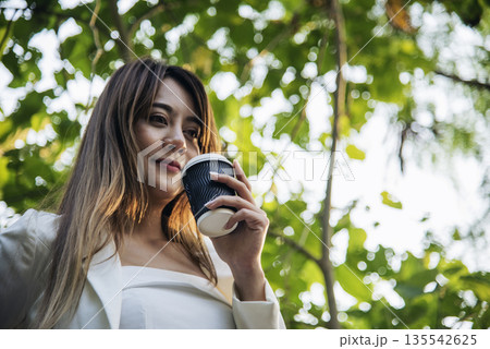 Woman drinking coffee hands holding hot disposable cup in green park. Happy Relax beautiful asian woman smiling face standing outdoors garden. Young women enjoy nature morning Freedom Lifestyle. Woman drinking coffee hands holding hot disposable cup in green park. Happy Relax beautiful asian woman smiling face standing outdoors garden. Young women enjoy nature morning Freedom Lifestyle. 135542625