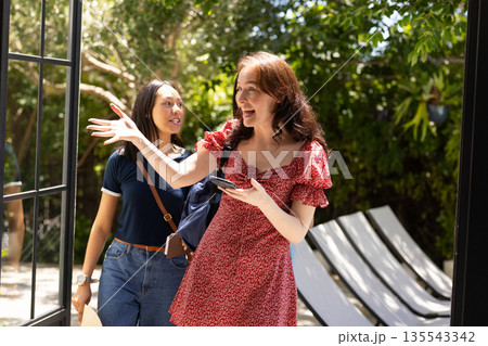 Two young women, one with long brown hair in red dress, happily entering sunlit patio Two young women, one with long brown hair in red dress, happily entering sunlit patio 135543342