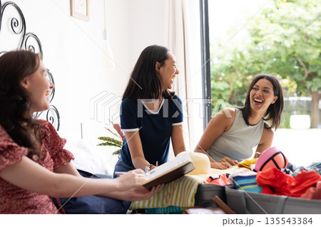 Laughing together, three young women with dark hair enjoy packing clothes in sunny room Laughing together, three young women with dark hair enjoy packing clothes in sunny room 135543384