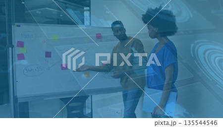 Gesturing coworker explaining whiteboard, other holding laptop listening, glass office partition 135544546